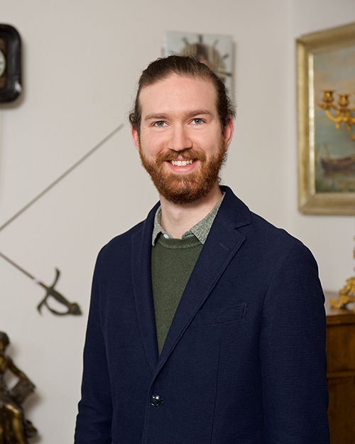 The man in charge of PR and social media wears a blue jacket, antiques can be seen in the background.