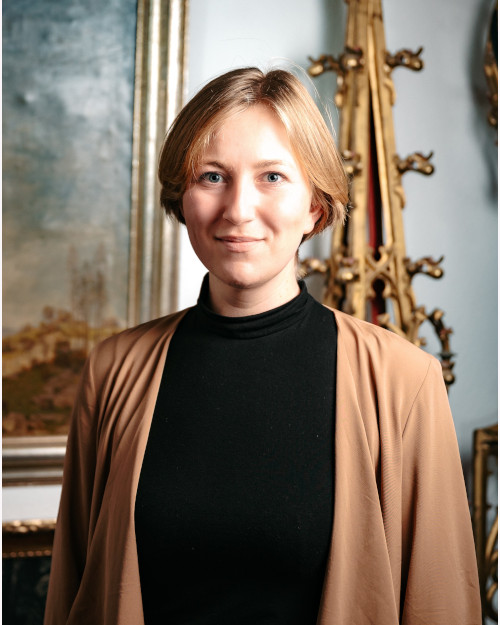 The art scholar is wearing a black sweater and a brown blazer. Antiques can be seen in the background.
